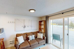 Ein Wohnzimmer mit braunem Sofa, Flachbildfernseher, großem Fenster und einer Schiebeglastür, die auf einen Balkon führt.