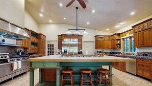 A spacious kitchen with wooden cabinets, a large island with stools, a stainless steel refrigerator, and a ceiling fan.
