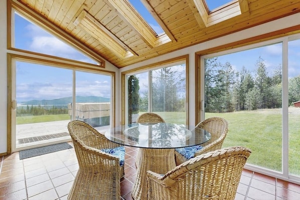 Ein Zimmer mit Glastisch, umgeben von Korbstühlen, großen Fenstern und einer Holzbalkendecke.