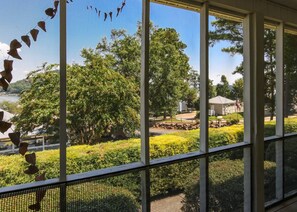 A view from a window with a metal railing, overlooking a well-maintained garden with various plants and trees, and a building with a flag in the background. - House #20 Sunshine Cottage (Not Pet Friendly) (Leesburg)