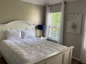 A bedroom with a white bed, a nightstand with a lamp, a window with curtains, and a wall-mounted artwork. - House #20 Sunshine Cottage (Not Pet Friendly) (Leesburg)
