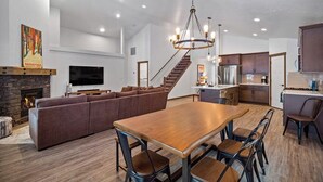 A spacious living area with a fireplace, a large sofa, a wooden dining table, and a kitchen in the background.