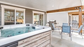 A hot tub with a wooden exterior, a patio area with chairs, and a grill.