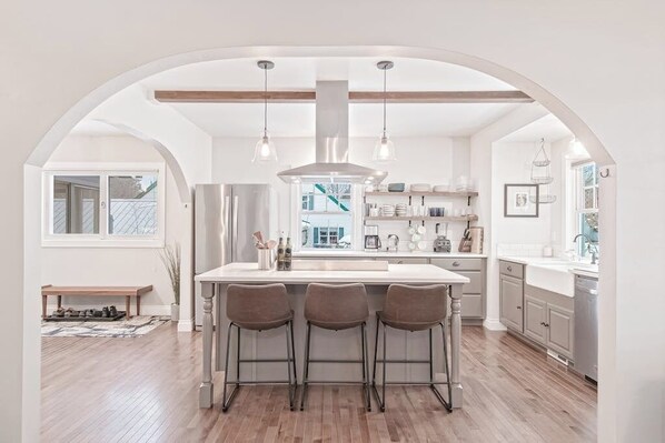 A kitchen with an island, bar stools, pendant lights, and a view through an archway. - Centrally-Located Upscale House with Free WiFi, Porch & Oversized Fenced Yard (Sandpoint)