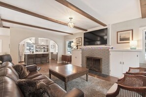 A spacious living room with a fireplace, a television, and a kitchen in the background. - Centrally-Located Upscale House with Free WiFi, Porch & Oversized Fenced Yard (Sandpoint)