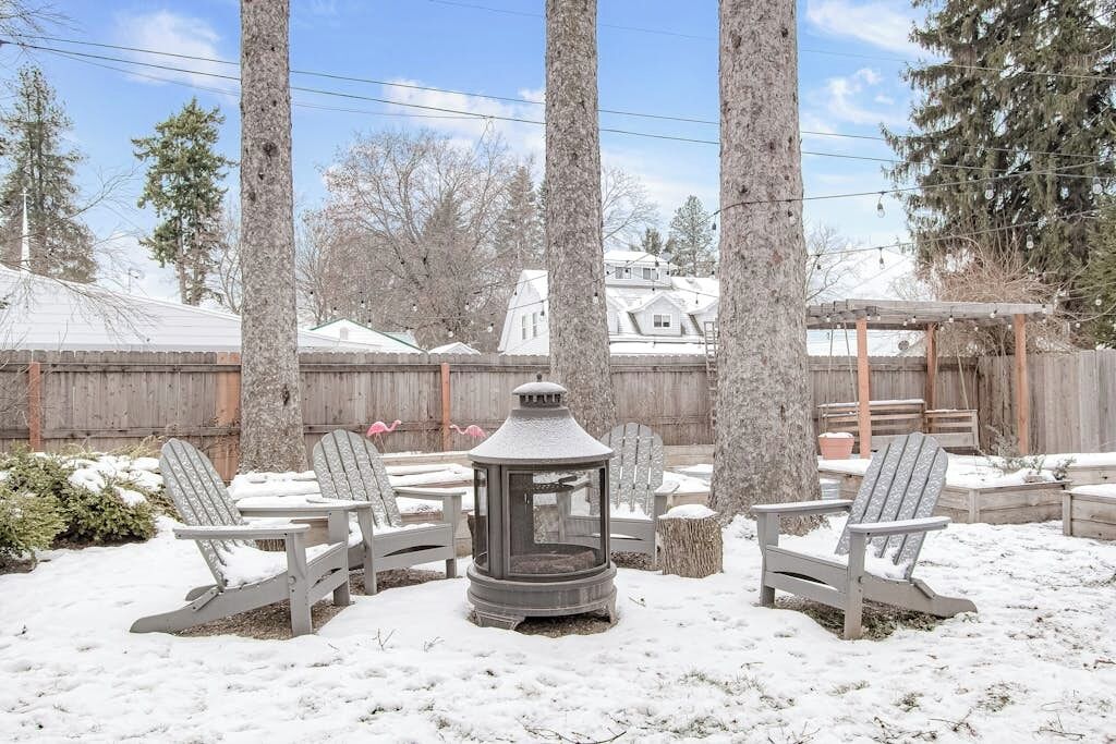 A snow-covered outdoor seating area with Adirondack chairs and a lantern, surrounded by trees and a wooden fence.