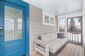 A blue door, a white sofa with cushions, a small wooden stool, and a window with a view of a snowy landscape. - Centrally-Located Upscale House with Free WiFi, Porch & Oversized Fenced Yard (Sandpoint)