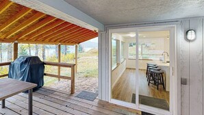 A covered outdoor area with a wooden deck, a barbecue grill, and a view of a kitchen with stools. - Charming home with river views, spacious patio, BBQ, WiFi and streaming (Sandpoint)