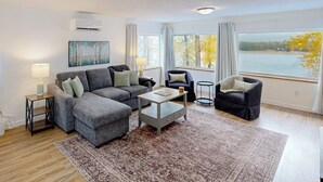 A modern living room with a grey sectional sofa, a coffee table, and a view of a lake through large windows. - Charming home with river views, spacious patio, BBQ, WiFi and streaming (Sandpoint)