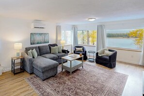 Ein modernes Wohnzimmer mit einem grauen Ecksofa, einem Couchtisch und Blick auf einen See durch große Fenster.