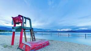 A lifeguard chair with a sign, a red lifeguard stand, and a sandy beach with a calm lake in the background. - Charming home with river views, spacious patio, BBQ, WiFi and streaming (Sandpoint)