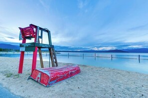 Ein Rettungsschwimmertisch mit Schild, ein roter Rettungsposten und ein sandiger Strand mit einem ruhigen See im Hintergrund.