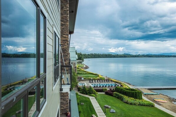 A lakeside view from a building with large windows, a swimming pool, and a well-maintained lawn. - Lake view condo with WiFi, balcony, fireplace, & shared pool/hot tub/grills (Sandpoint)