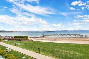 A waterfront property with a grassy lawn, a dock extending into the water, and mountains in the background. - Lake view condo with WiFi, balcony, fireplace, & shared pool/hot tub/grills (Sandpoint)