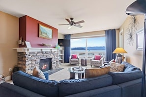 A living room with a stone fireplace, a ceiling fan, a sofa, and a view of the ocean. - Lake view condo with WiFi, balcony, fireplace, & shared pool/hot tub/grills (Sandpoint)
