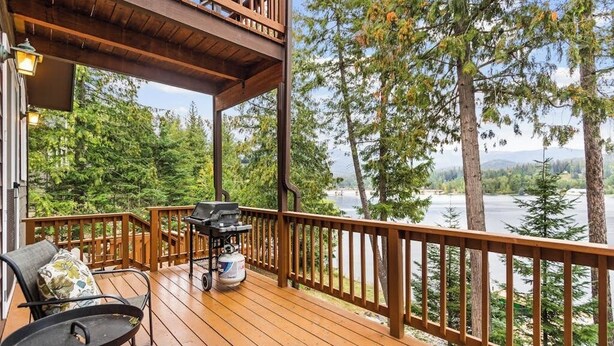 A wooden deck with a grill and chairs, overlooking a lake and forest. - Riverfront home with mountain view, hot tub, deck, WiFi, W/D, & fireplace (Sagle)