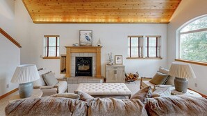 A living room with a fireplace, wooden ceiling, and large windows. - Riverfront home with mountain view, hot tub, deck, WiFi, W/D, & fireplace (Sagle)