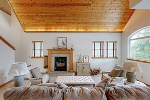 A living room with a fireplace, wooden ceiling, and large windows. - Riverfront home with mountain view, hot tub, deck, WiFi, W/D, & fireplace (Sagle)