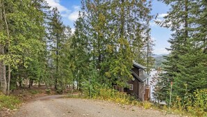 A cabin with a gravel driveway, surrounded by dense forest and a clear sky. - Riverfront home with mountain view, hot tub, deck, WiFi, W/D, & fireplace (Sagle)
