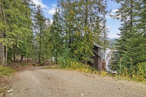 A cabin with a gravel driveway, surrounded by dense forest and a clear sky. - Riverfront home with mountain view, hot tub, deck, WiFi, W/D, & fireplace (Sagle)