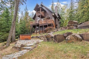 A cabin with a deck, surrounded by trees and rocks. - Riverfront home with mountain view, hot tub, deck, WiFi, W/D, & fireplace (Sagle)