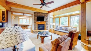 A spacious living room with a stone fireplace, a ceiling fan, and large windows. - Lakefront home with mtn views, private hot tub, dock, deck, & grill (Sandpoint)