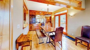 A wooden dining area with benches and chairs, a kitchen in the background, and a large window with a view. - Lakefront home with mtn views, private hot tub, dock, deck, & grill (Sandpoint)