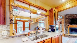 A kitchen with wooden cabinets, a stone fireplace, and a large window. - Lakefront home with mtn views, private hot tub, dock, deck, & grill (Sandpoint)