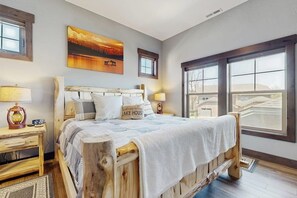 A bedroom with a wooden bed, a nightstand with a lamp, a painting on the wall, and a large window. - Beautiful condo with river views, deck, BBQ, WiFi, and nearby activities (Sandpoint)