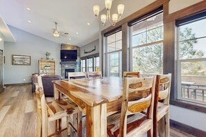A spacious dining area with a large wooden table and chairs, large windows with a view of trees, and a modern kitchen in the background. - Beautiful condo with river views, deck, BBQ, WiFi, and nearby activities (Sandpoint)