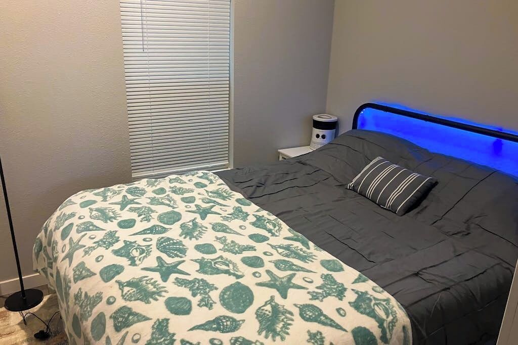 A bedroom with a bed, a bedside table, a lamp, and a window with blinds.