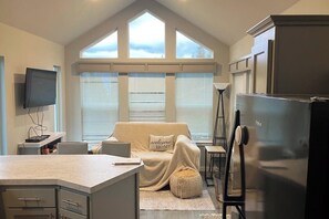 Una cocina con refrigerador empotrado, comedor con mesa y sillas, sofá con cojín que dice 'bienvenido a casa', y ventanas amplias con persianas.
