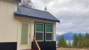 Une petite cabane avec une porte, deux fenêtres, un escalier en bois et une vue sur les montagnes et les arbres.