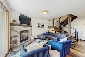 Ein Wohnzimmer mit Kamin aus Stein, einem Ecksofa, einem an der Wand montierten Fernseher und einer Treppe.