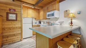 A kitchen with wooden cabinets, a white refrigerator, a microwave, a sink, and a bar area with stools. - Convenient Studio, Walking Distance to Sun Valley Lake - Shopping, Skiing Nearby (Sun Valley)