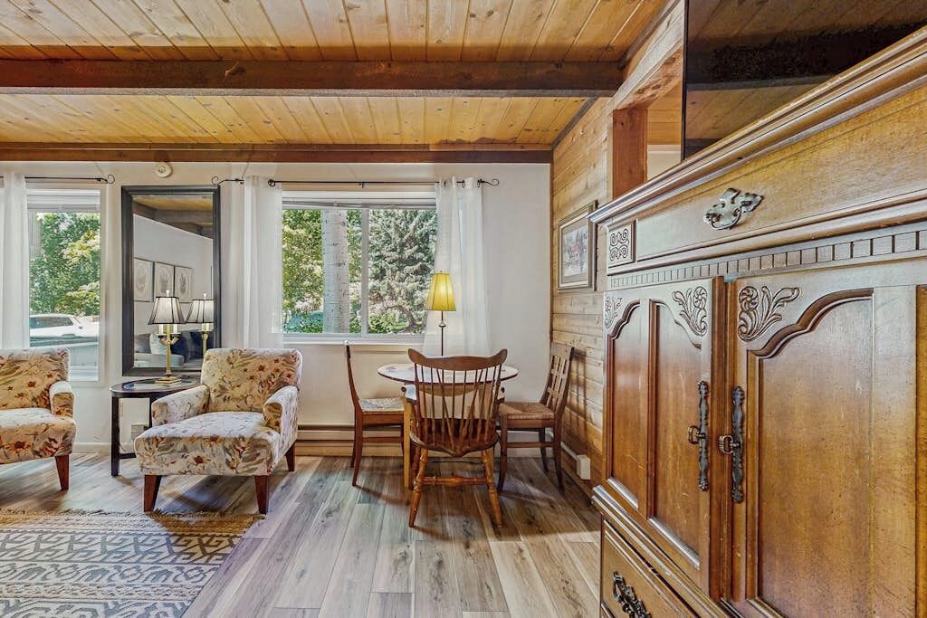 A room with wooden floors, a dining table with chairs, a cabinet with ornate handles, and a window with curtains.
