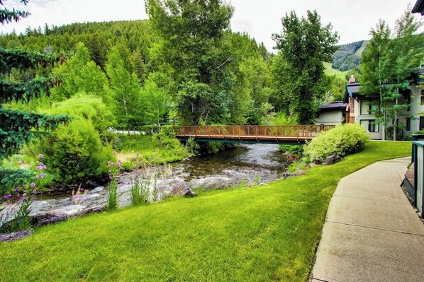 A riverside view with a wooden bridge, lush greenery, and a building with a sloped roof.