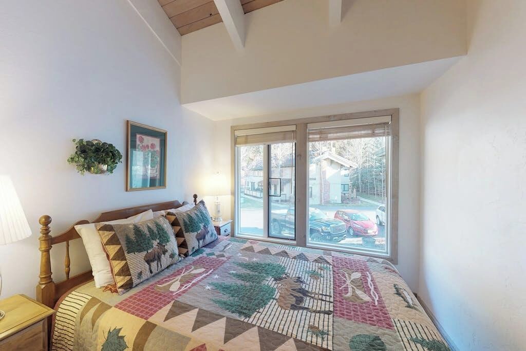 Quarto com uma janela grande, cama com colcha estampada, mesa de cabeceira e planta fixada na parede.