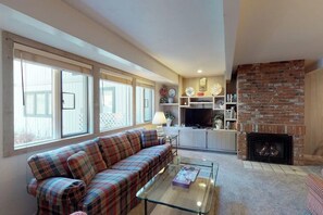 A living room with a sofa, a glass coffee table, a fireplace, and a kitchen area with a TV and shelves.