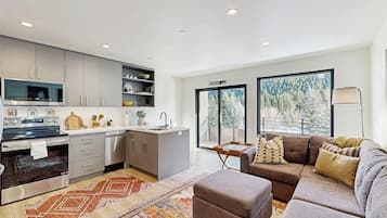 Una cocina moderna equipada con horno, microondas y lavavajillas, zona de comedor con sofá y alfombra, y ventanas amplias con vista a árboles.