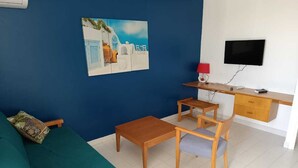 Ein Zimmer mit einem türkisfarbenen Sofa, einem Holztisch mit zwei Stühlen, einem Schreibtisch mit Fernseher und einem Bild an der Wand.