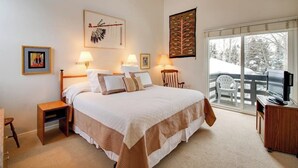 A bedroom with a large bed, two bedside tables, a chair, a desk, a television, and a balcony with a view of trees. - Cozy Condo w/ Free WiFi, Heated Pool & Tennis - Close to Skiing (Sun Valley)