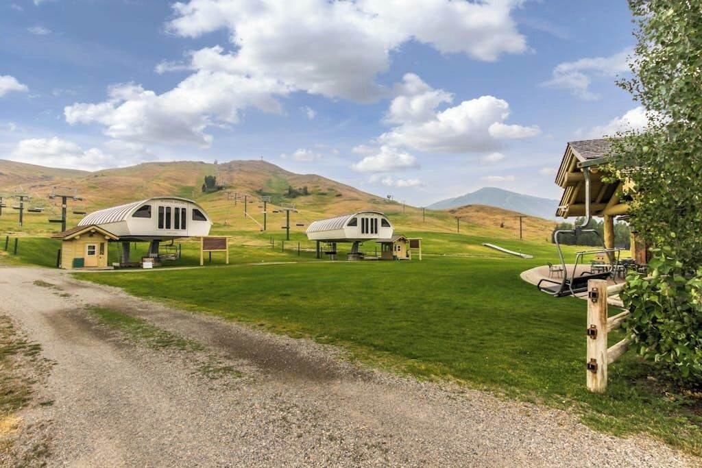 Un cadre rural avec deux cabanes blanches à un seul étage, dotées de vérandas attenantes, et entourées de champs et de collines verdoyantes.
