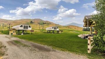 Un cadre rural avec deux cabanes blanches à un seul étage, dotées de vérandas attenantes, et entourées de champs et de collines verdoyantes.