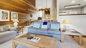 A living room with a sofa, coffee table, and lamps. There is a wooden ceiling and a kitchen area in the background. - Rustic home with gas fireplace, pool & hot tub - ski-in/out to Dollar Mountain (Sun Valley)
