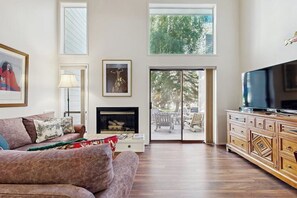 A living room with a fireplace, a television, and a large window overlooking a deck. - Views and recreation: Sun Valley condo with pool, sauna, tennis and hot tub! (Sun Valley)