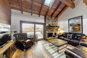 A cozy living room with a stone fireplace, leather sofas, a large window, and a framed picture of a mountain. - Ski-in/ski-out alpine condo with prime location, shared pool, hot tub, & tennis (Sun Valley)
