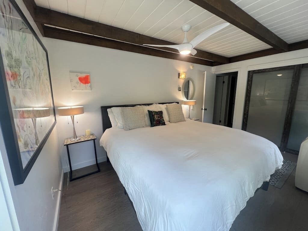 Un dormitorio con una cama grande, un ventilador de techo, una mesita de noche con lámpara y una puerta que da a otra habitación.