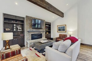Ein Wohnzimmer mit Kamin aus Stein, einem darüber montierten Fernseher, einer Couch, einem gemusterten Teppich und einem Holztisch.