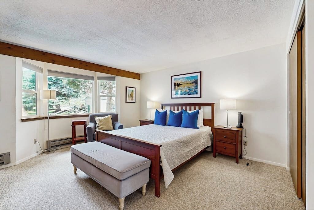 A bedroom with a large bed, a bench, a nightstand, a lamp, and a window with a view of greenery.